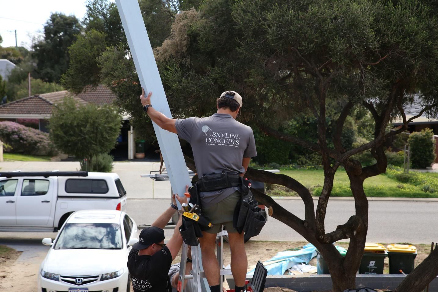 Worker on a truck lifts a long white beam near a suburban street, with trees and parked cars nearby
