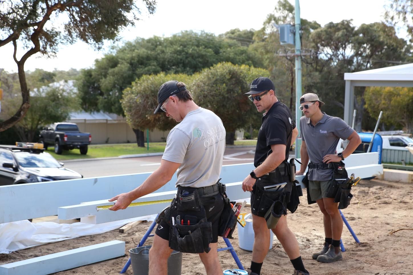 Three workers in tool belts handle a long white board beside a truck at a roadside worksite.