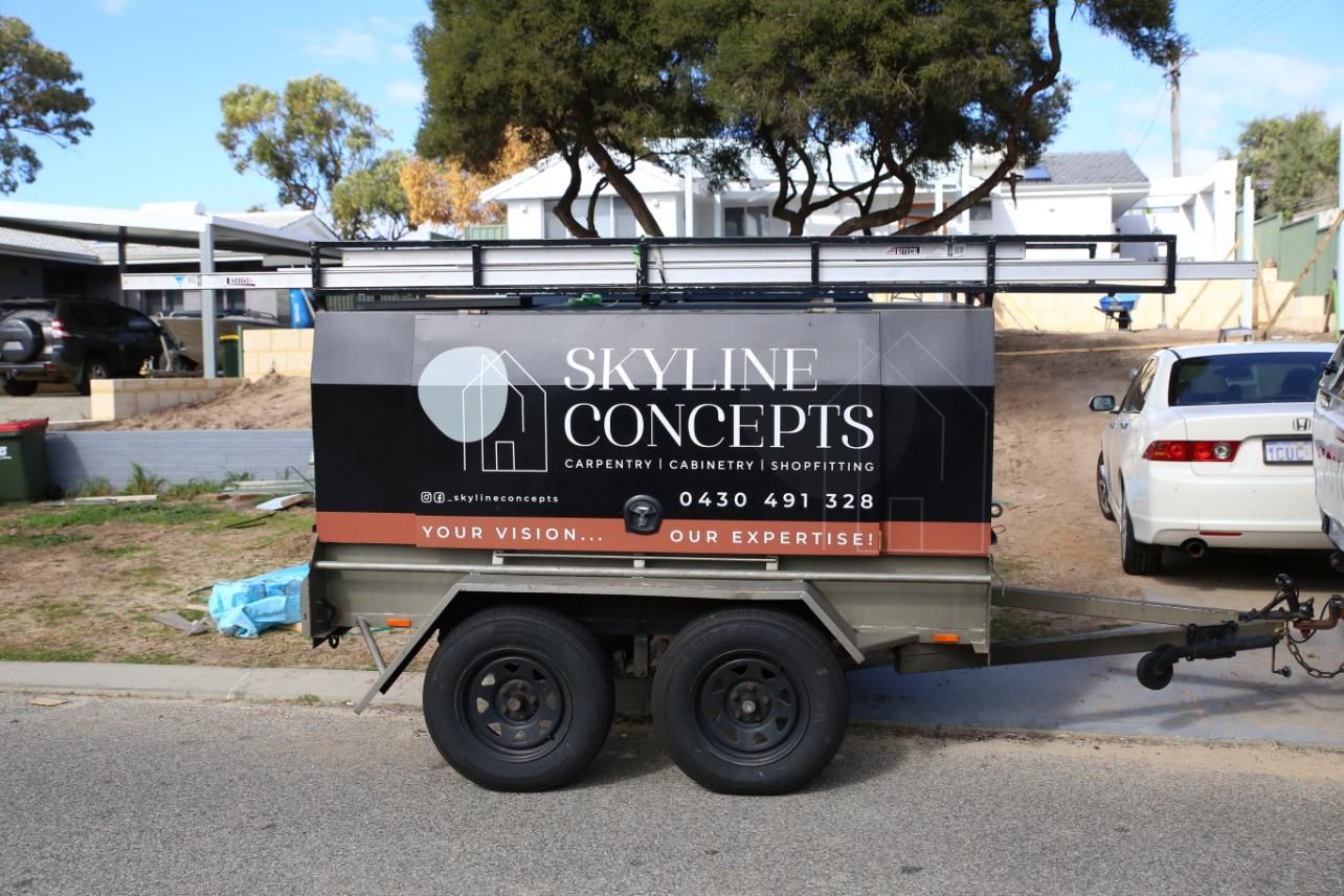 Black trailer with “Skyline Concepts” parked on a street beside a white car and trees