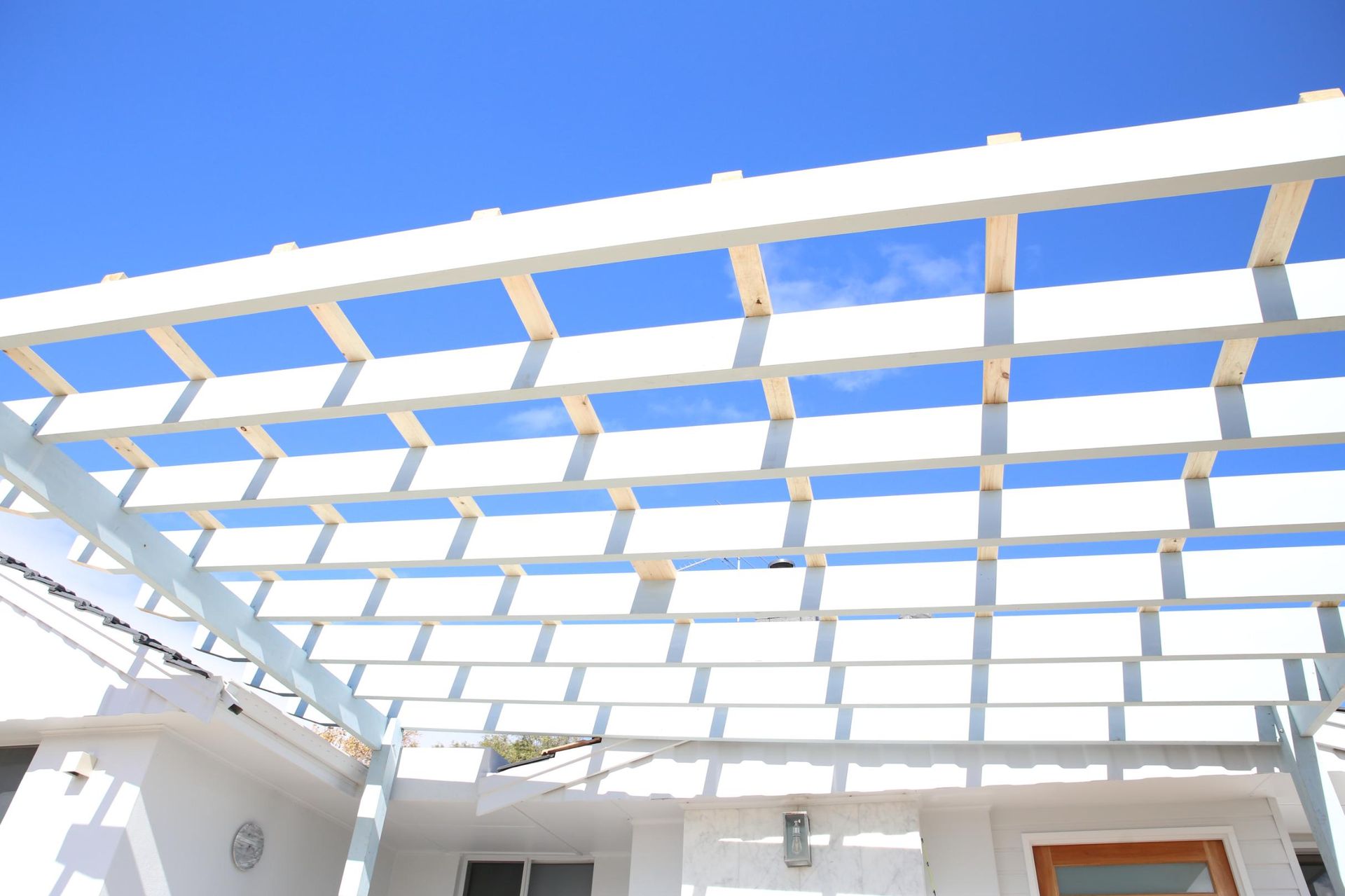 White pergola roof grid against a bright blue sky over a modern building courtyard