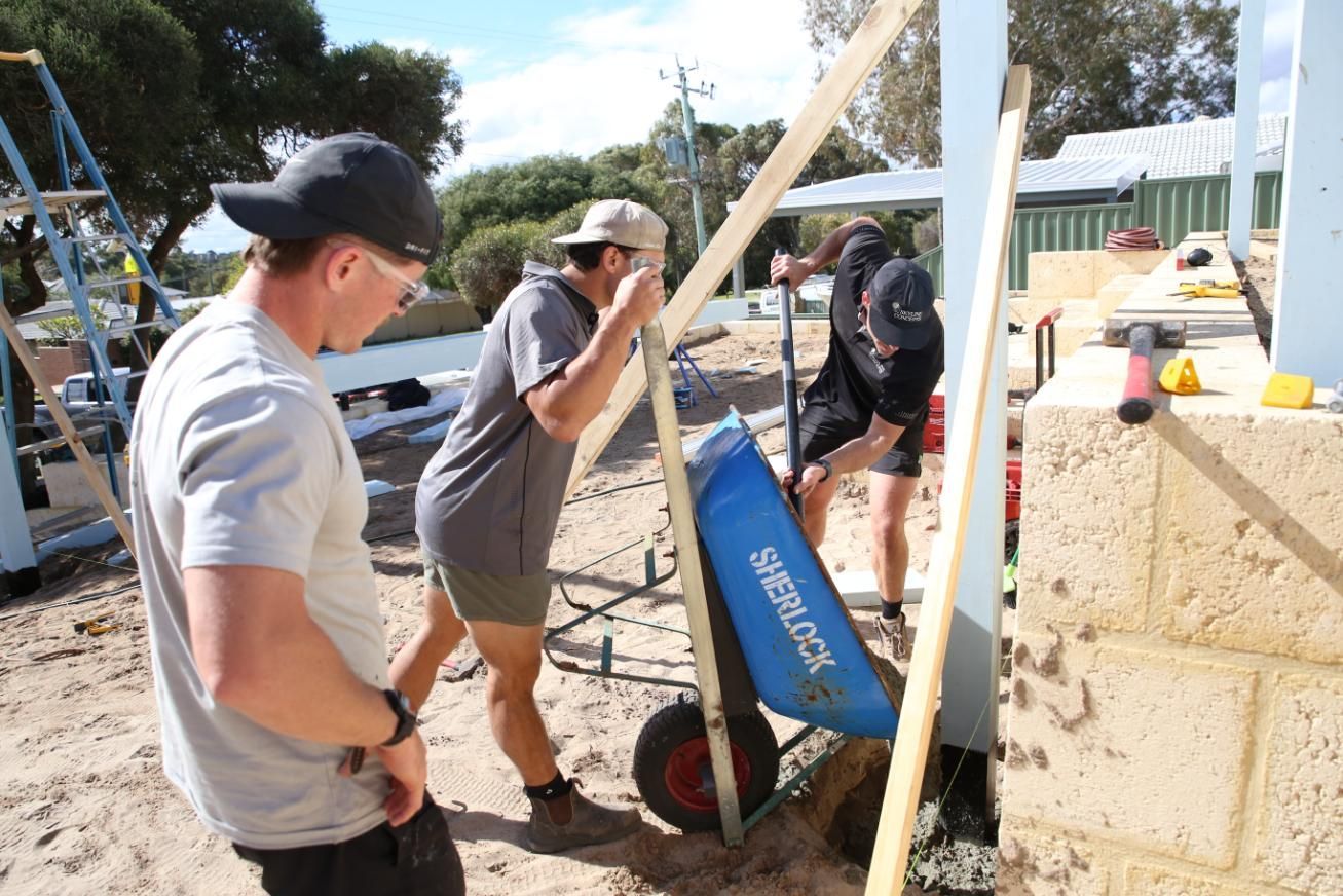 Workers operating a blue machine at an outdoor construction site beside a concrete wall