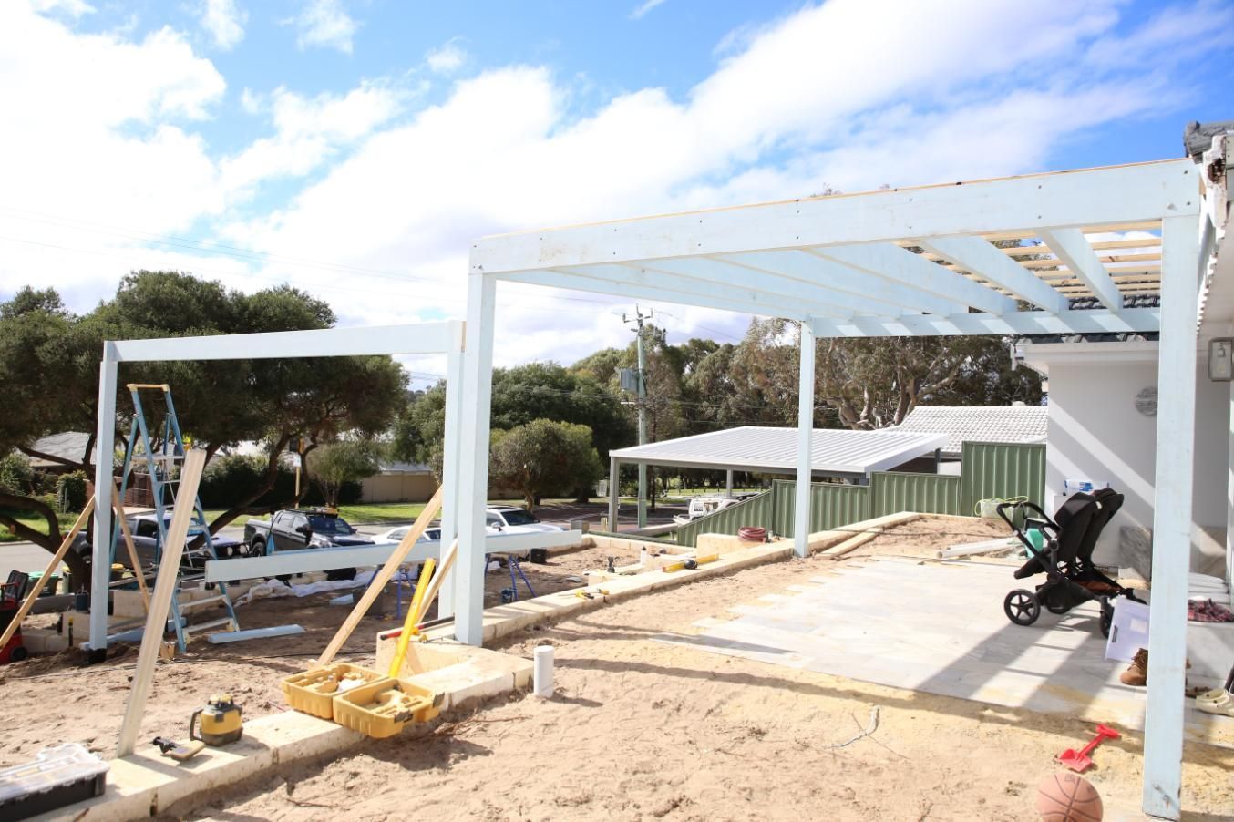 Construction site with white steel pergola frame, tools, and wheelbarrow on a sandy lot under a blue sky