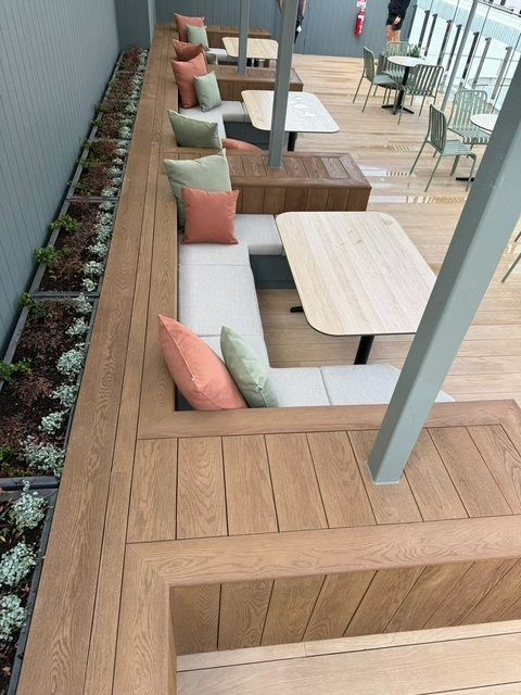 Outdoor café seating with wooden benches, light tables, and pastel cushions under a covered walkway