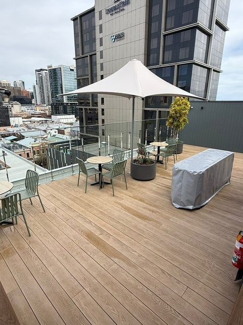 Rooftop deck with wooden flooring, tables, chairs, a white umbrella, and city buildings in the background