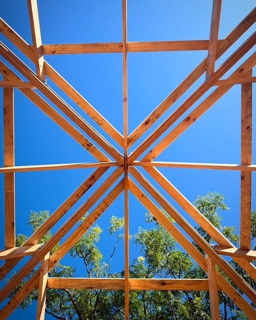Wooden roof frame with diagonal beams against a bright blue sky and trees below