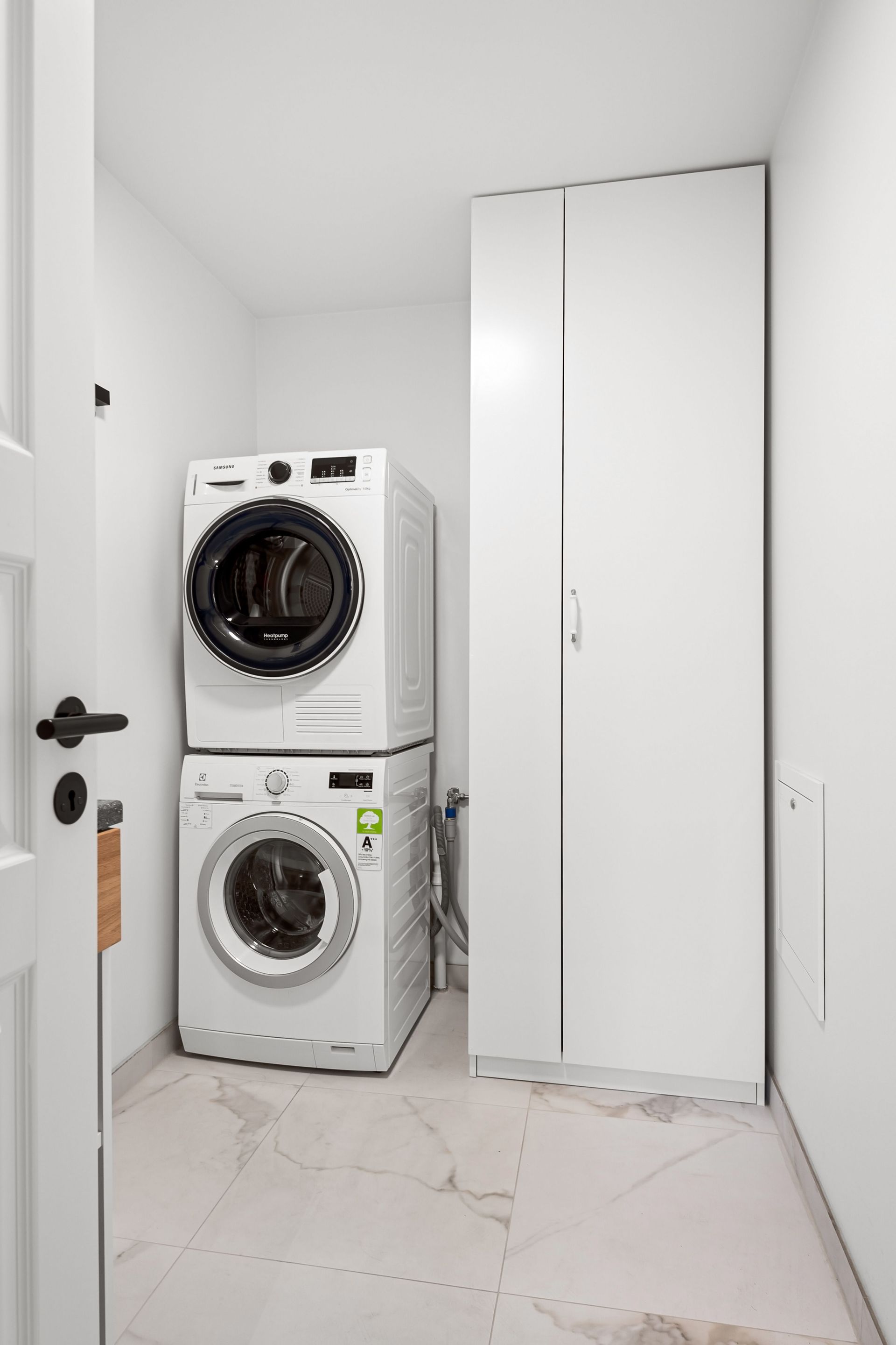 Stacked washer and dryer beside a tall white storage cabinet in a narrow laundry room.