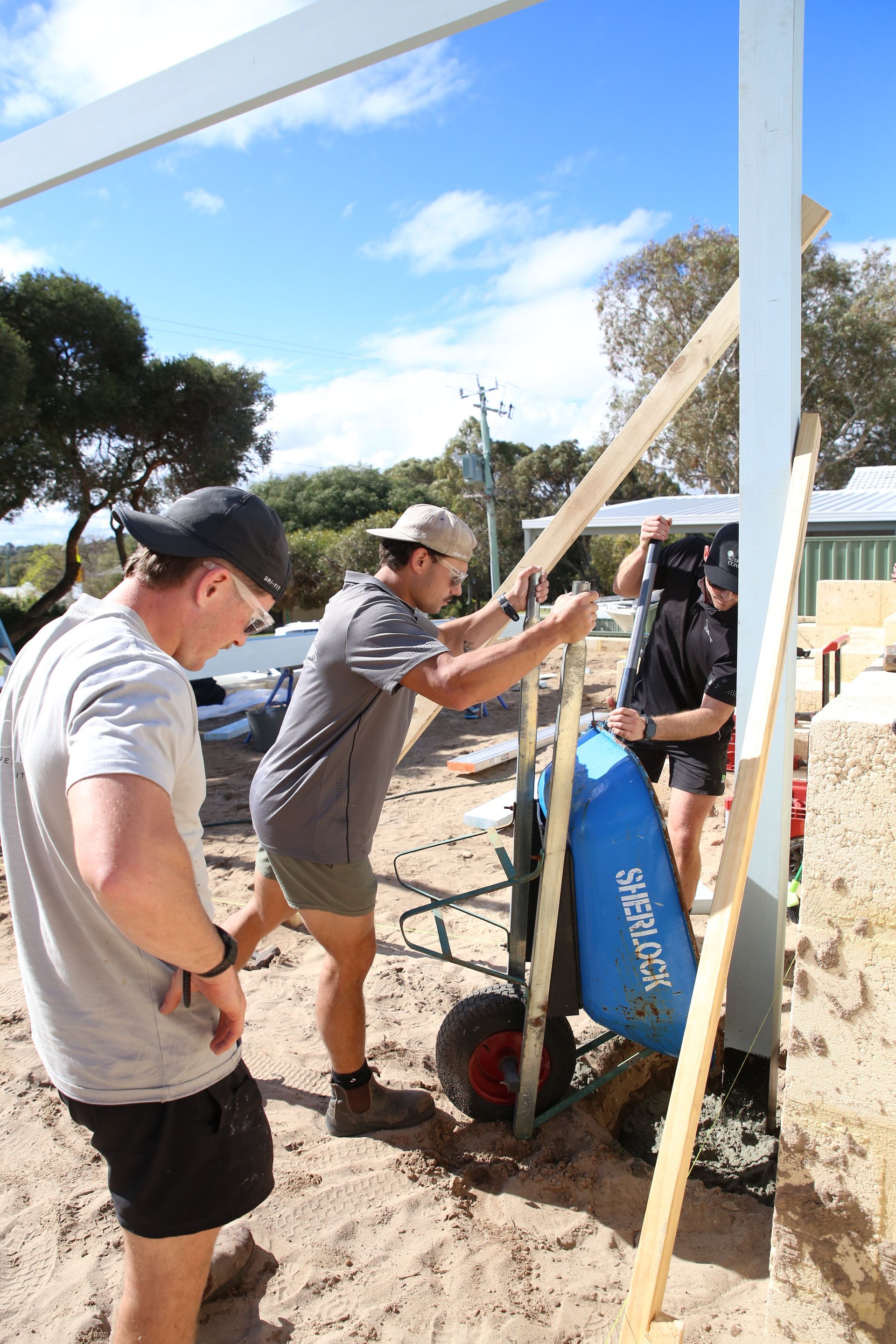 Three people assembling a wooden post at a sandy outdoor event with blue sky overhead