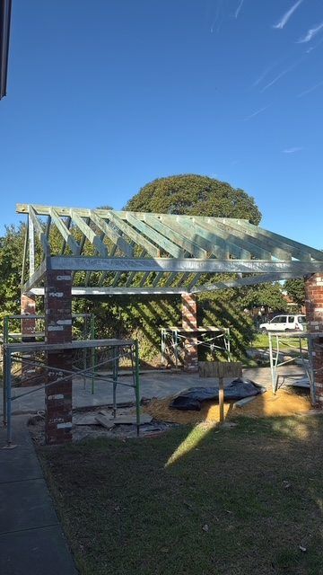 Small greenhouse with a slanted glass roof in a sunny yard, framed by a tree and blue sky.
