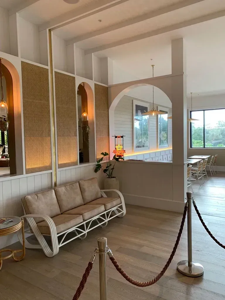 Elegant lounge with beige sofa, arched mirrors, woven wall panels, and large windows overlooking a bright waterfront.