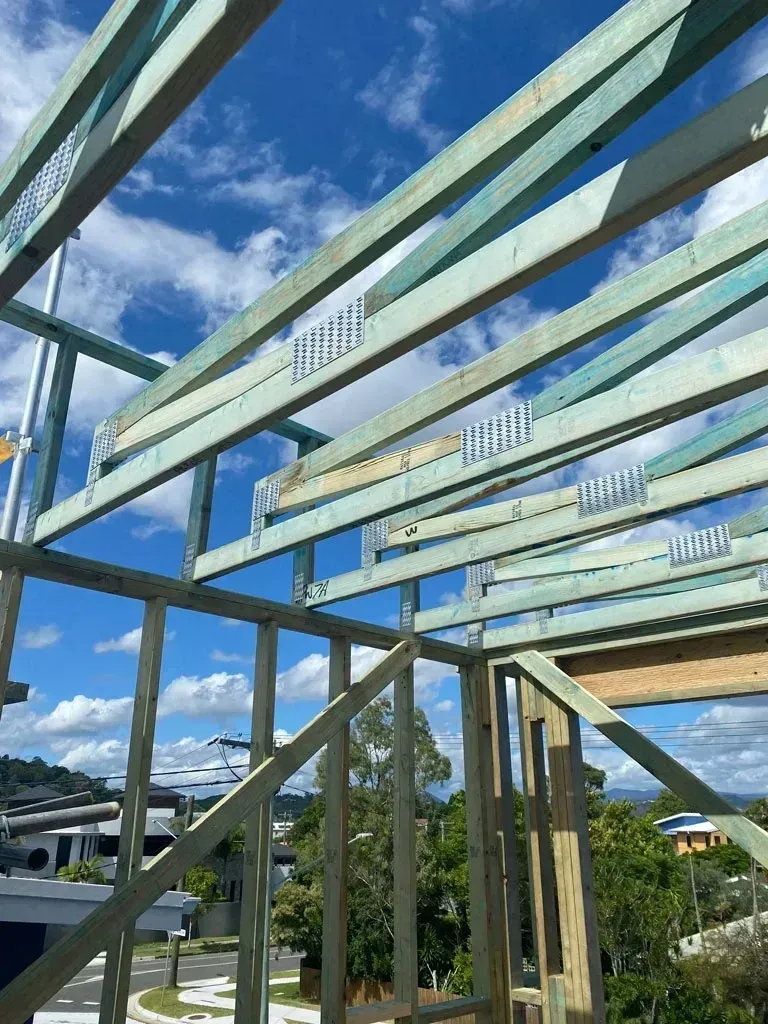 Steel frame under construction against blue sky and clouds, with trees in the background.