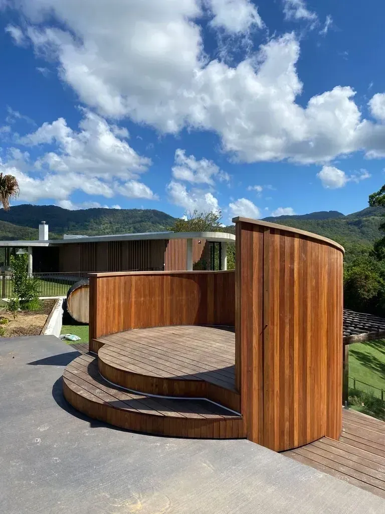 Round wooden outdoor pod with curved walls and circular bench, set on a paved path under a blue sky.