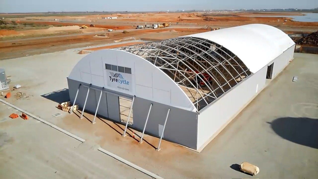 Large white arched warehouse under construction in a sandy industrial area, roof partially exposed.