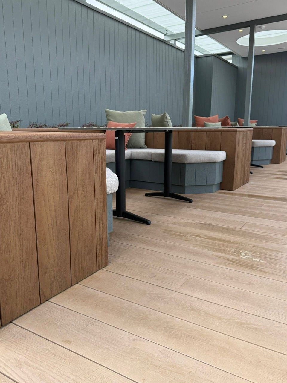 Covered outdoor lounge with wooden planters, gray seating, and a light wood deck under a roof.