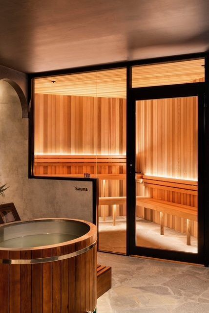 Wood-paneled sauna with a round hot tub and glass door, lit by warm amber light.