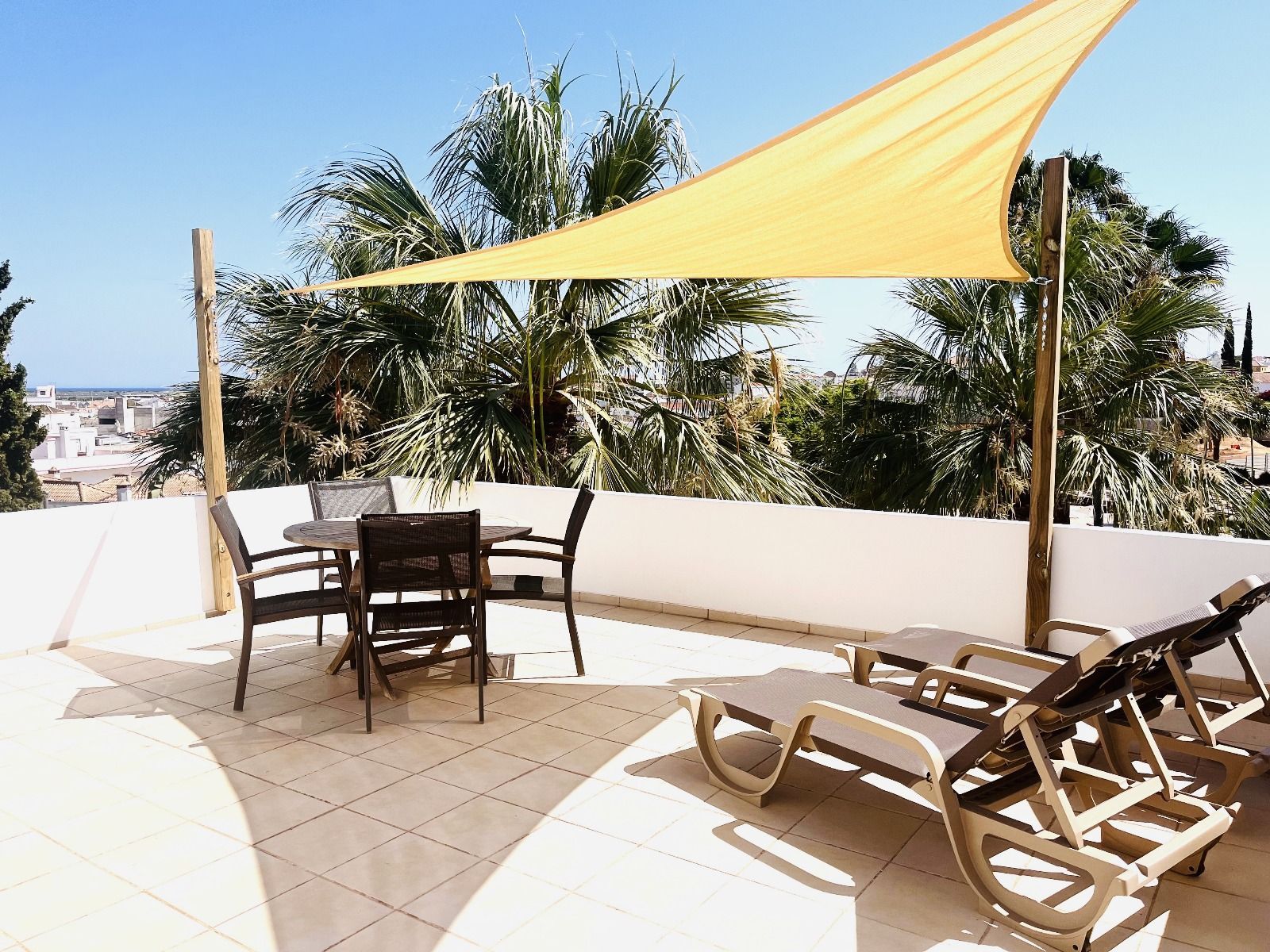 Terras met tafel, stoelen en ligstoelen onder een gele parasol. Palmbomen en uitzicht op de stad op de achtergrond.