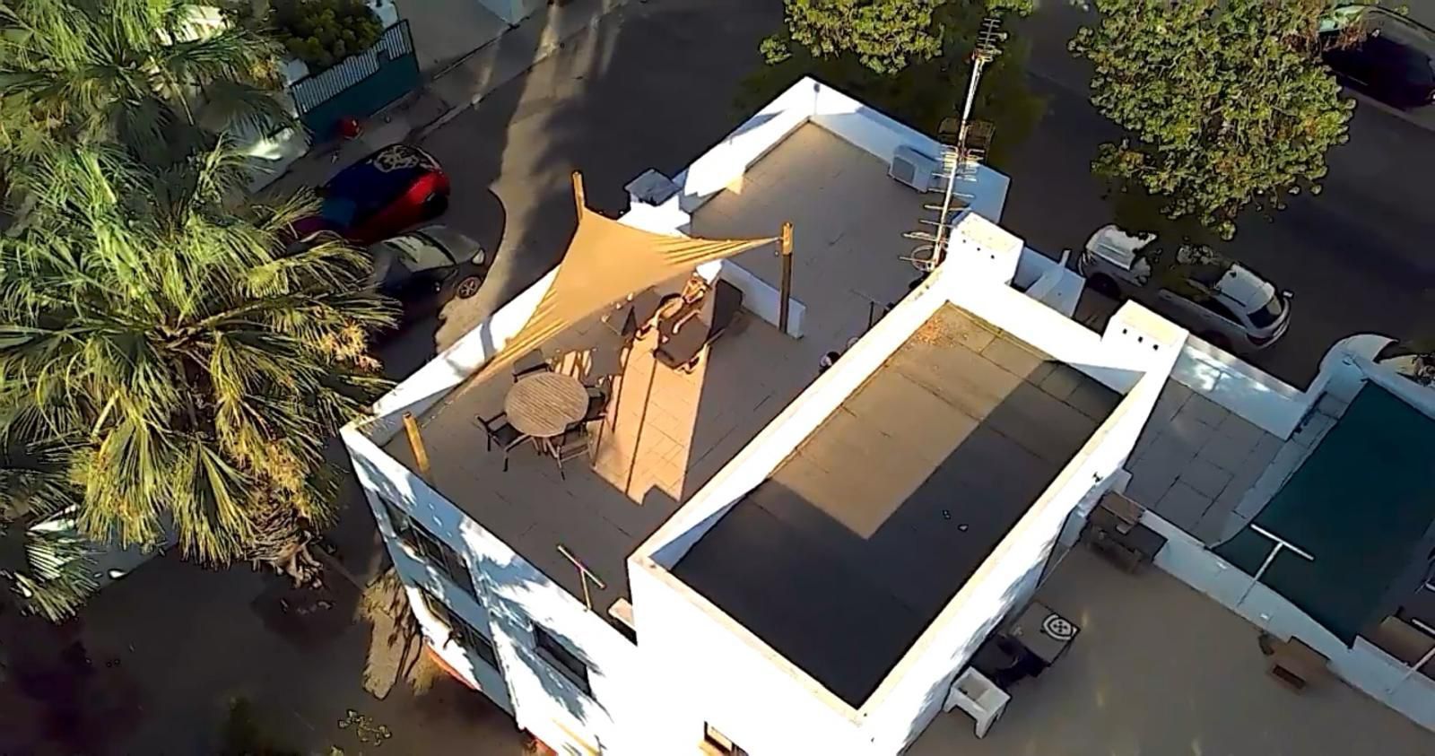 Luchtfoto van een wit gebouw met een plat dak, tafels, stoelen en een parasol. Er zijn een straat en geparkeerde auto's zichtbaar.