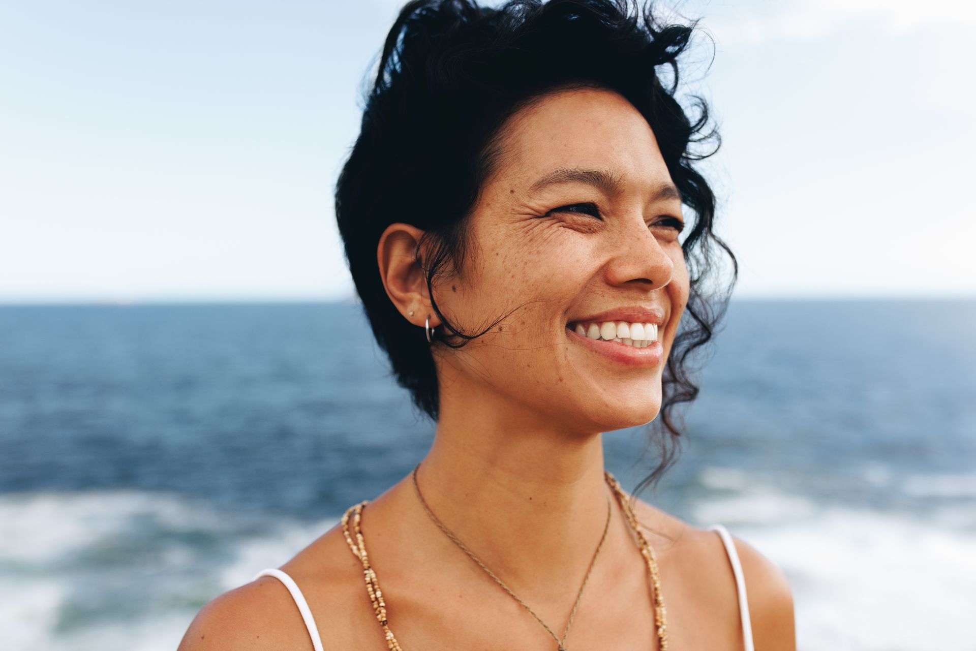 Smiling person with curly hair by the ocean, wearing a white top and necklace