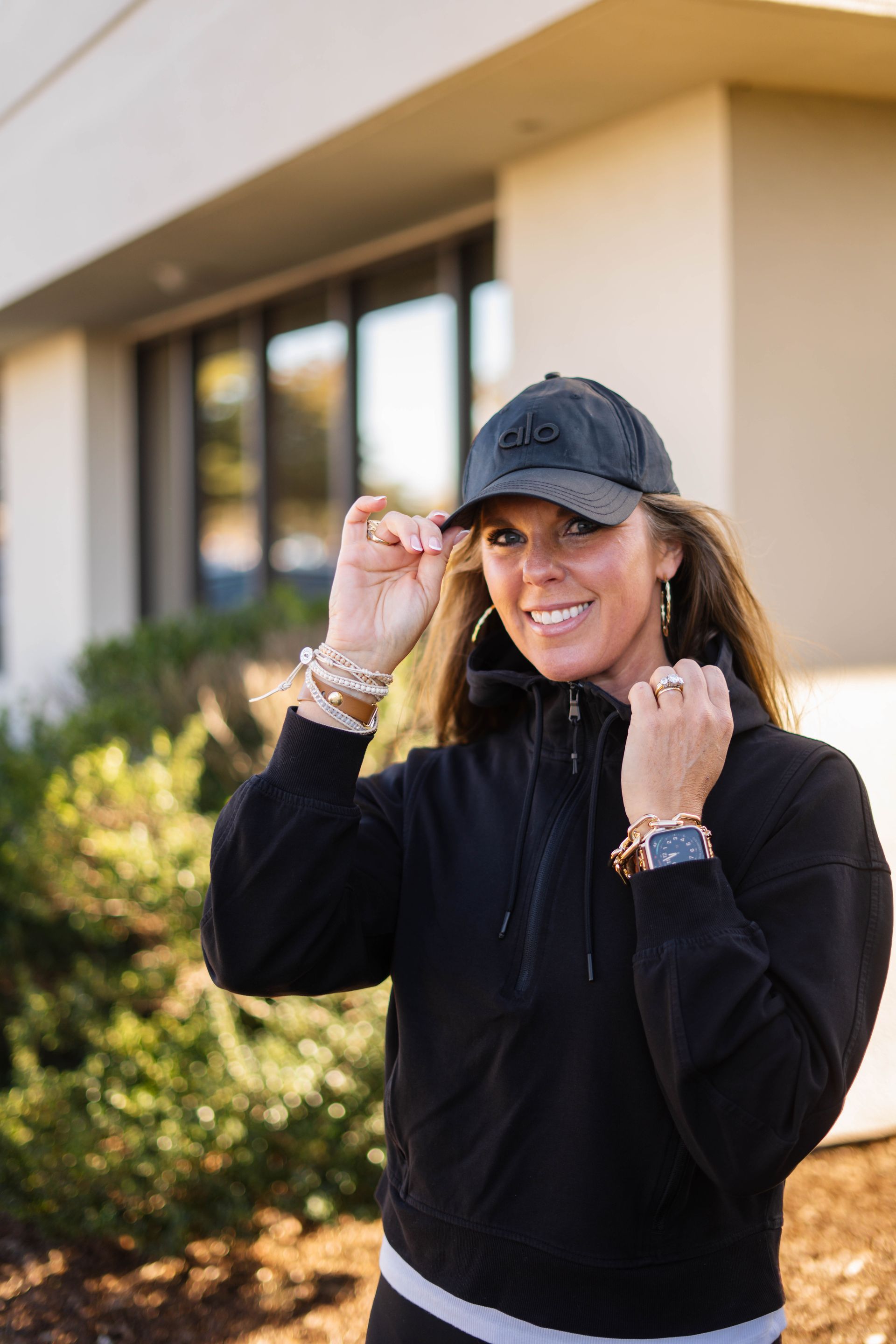 Smiling person in a black cap and sweater standing outdoors near a building, adjusting their cap and collar.