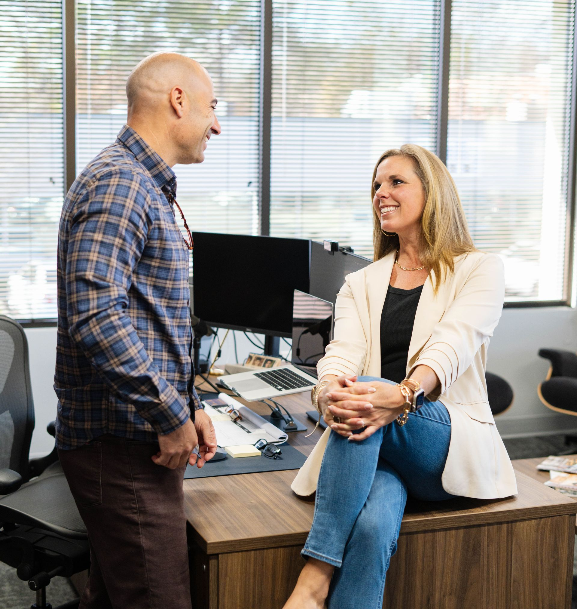 Two coworkers talking in an office, one seated on a desk and smiling
