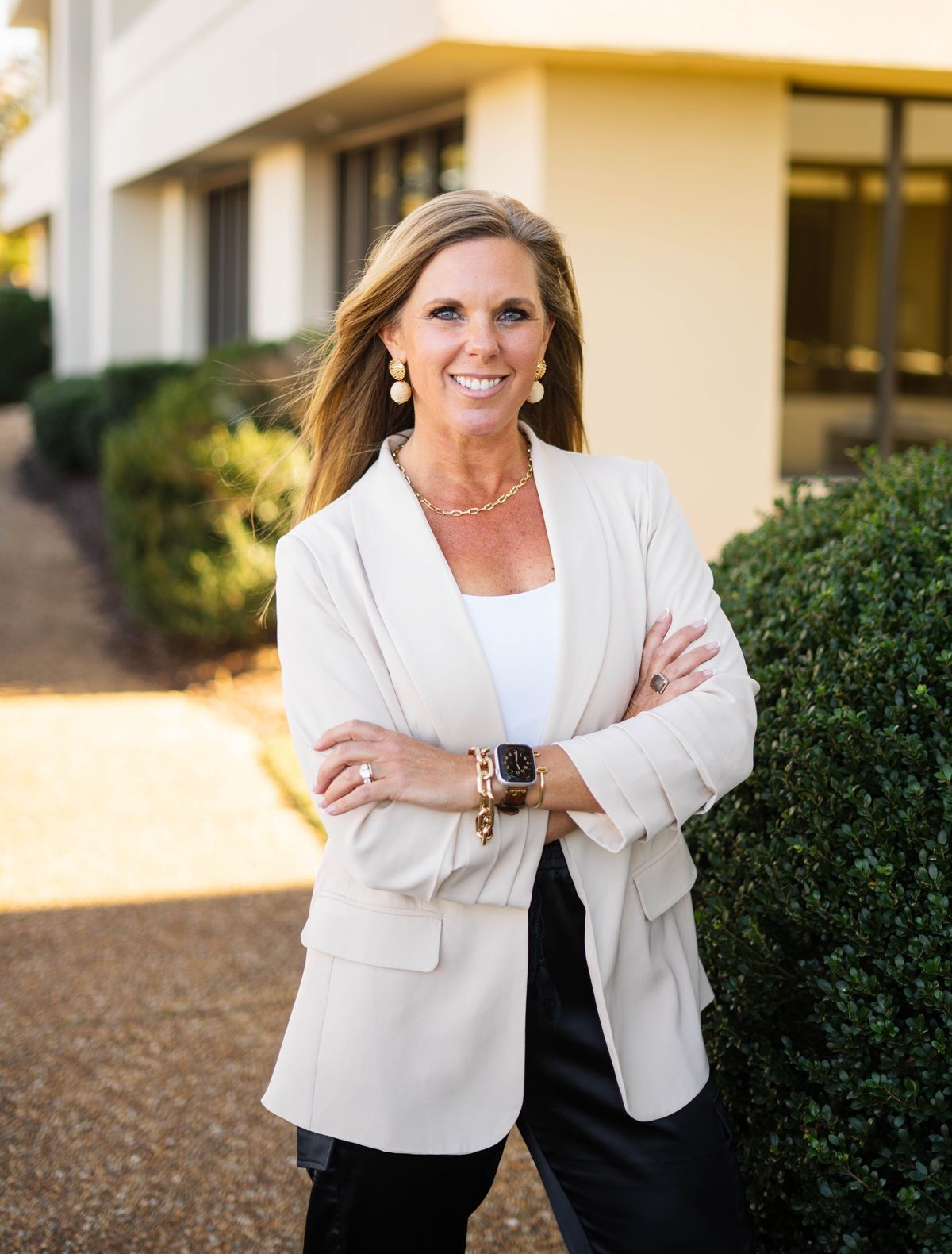Woman in a white blazer standing outdoors beside a building, smiling with arms crossed.