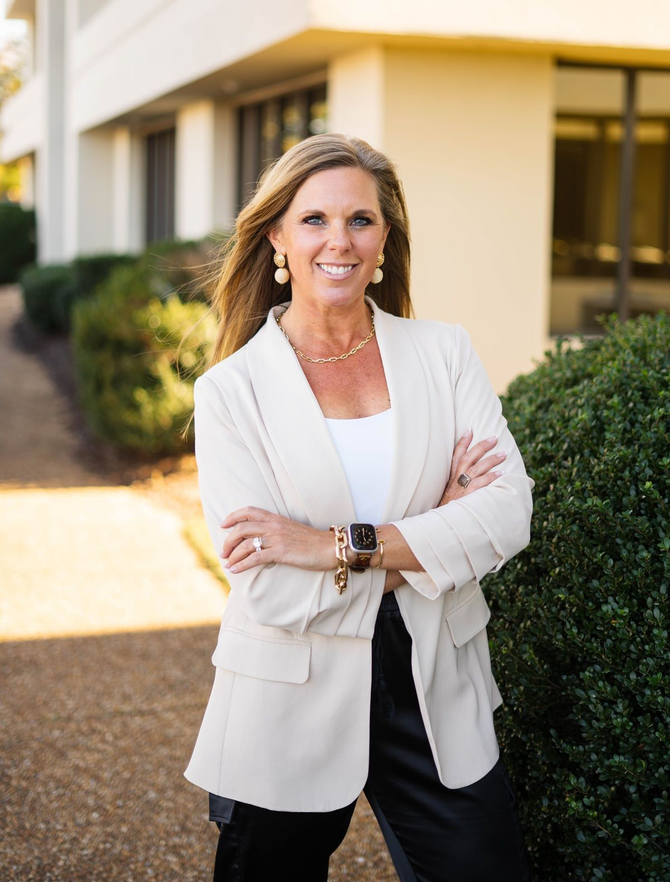 Woman in a white blazer standing outdoors beside a building, smiling with arms crossed.