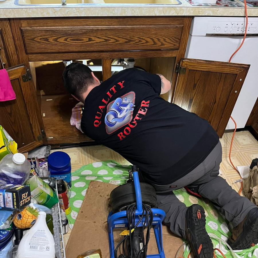 Person kneeling under kitchen sink cabinet, working on plumbing repairs with tools scattered nearby.