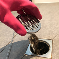 Gloved hand lifting hair clump from a bathroom drain with a metal drain cover nearby