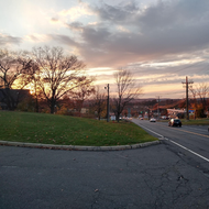 Sunset over a quiet suburban street with bare trees, grassy hill, and a parked car.