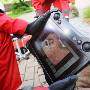 Gloved hands holding a rugged inspection tablet showing a close-up of a damaged pipe or wall cavity