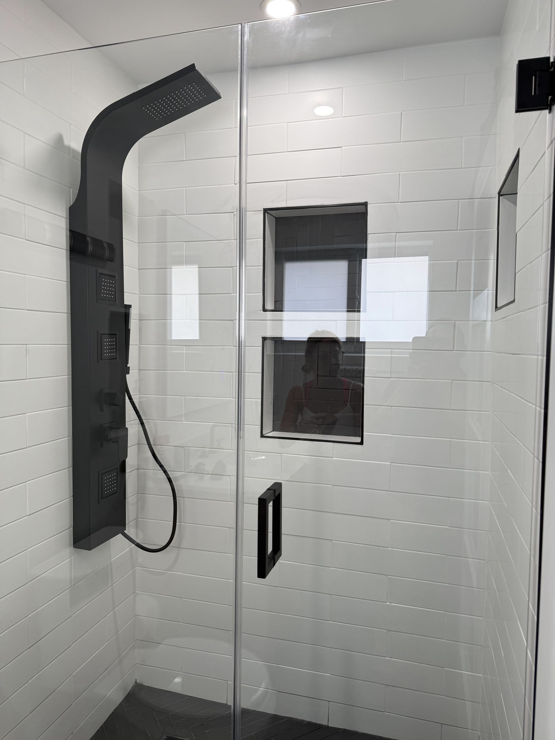 Modern shower with black fixtures, glass door, white tile, and a person reflected in the glass.