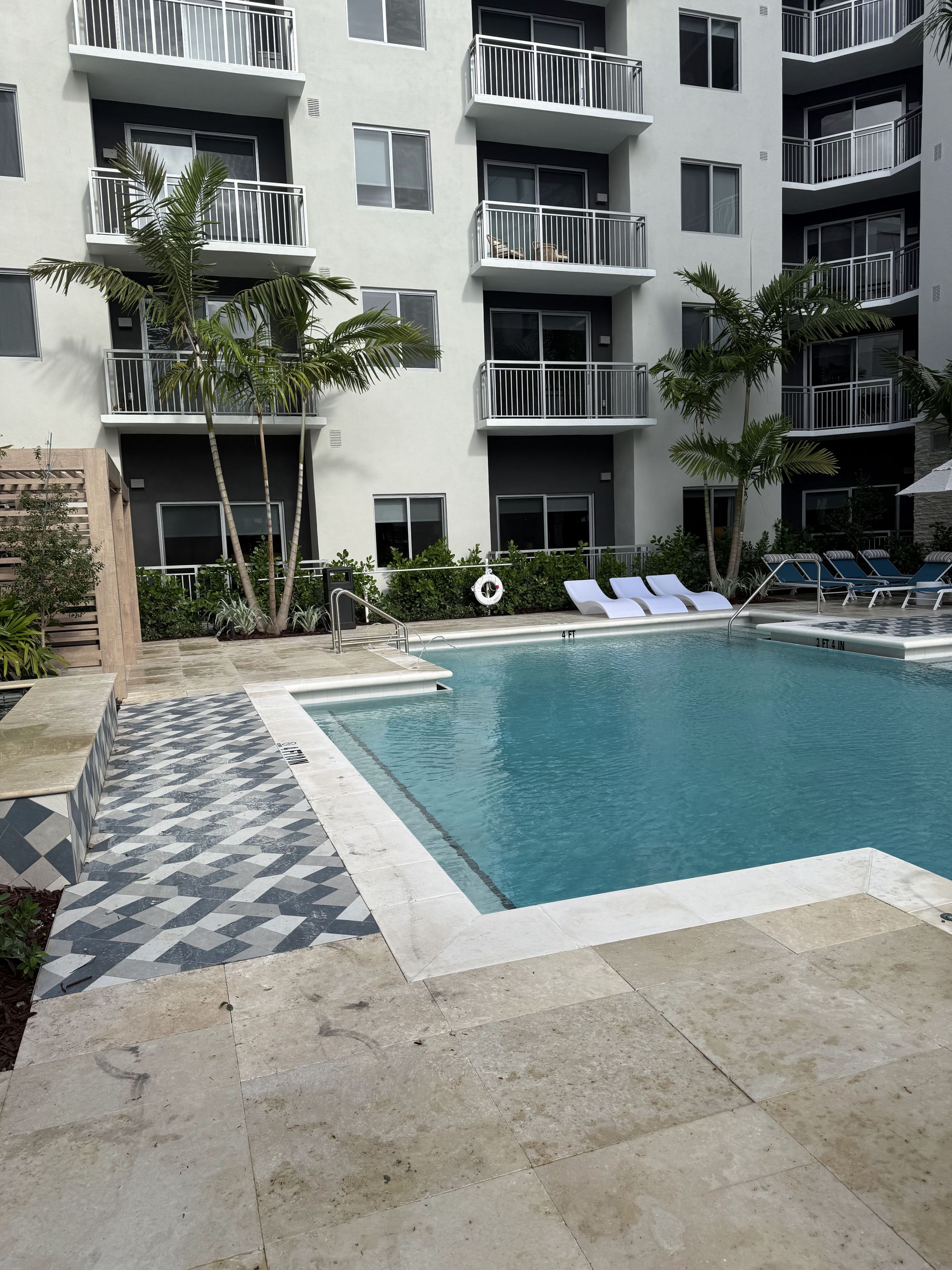 Swimming pool in front of a white apartment building with balconies. Lounge chairs and trees surround the pool.