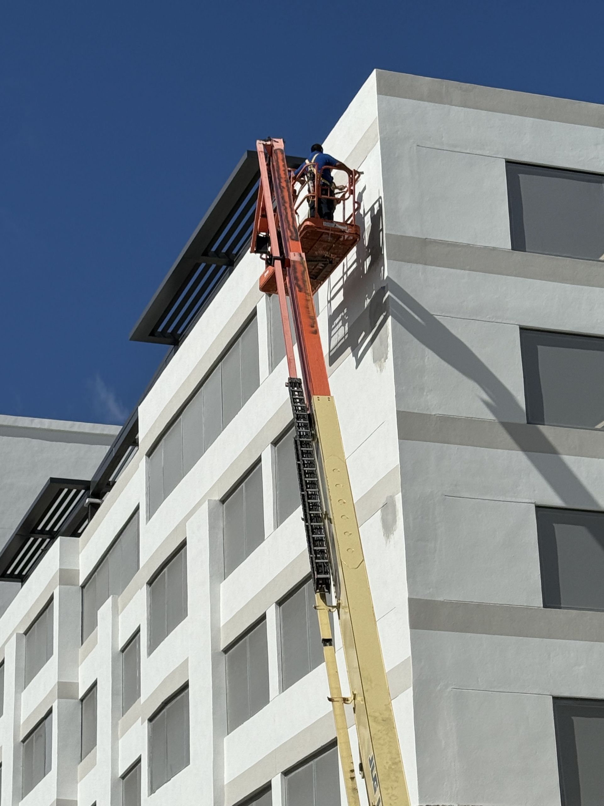 Person in orange lift working on the exterior of a gray building under a blue sky.