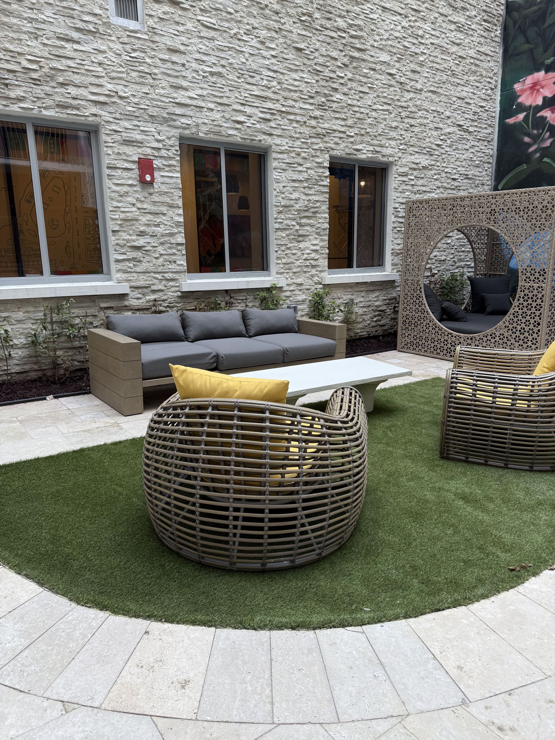 Outdoor seating area with grey sofa, woven chairs, and artificial grass. Stone wall background.