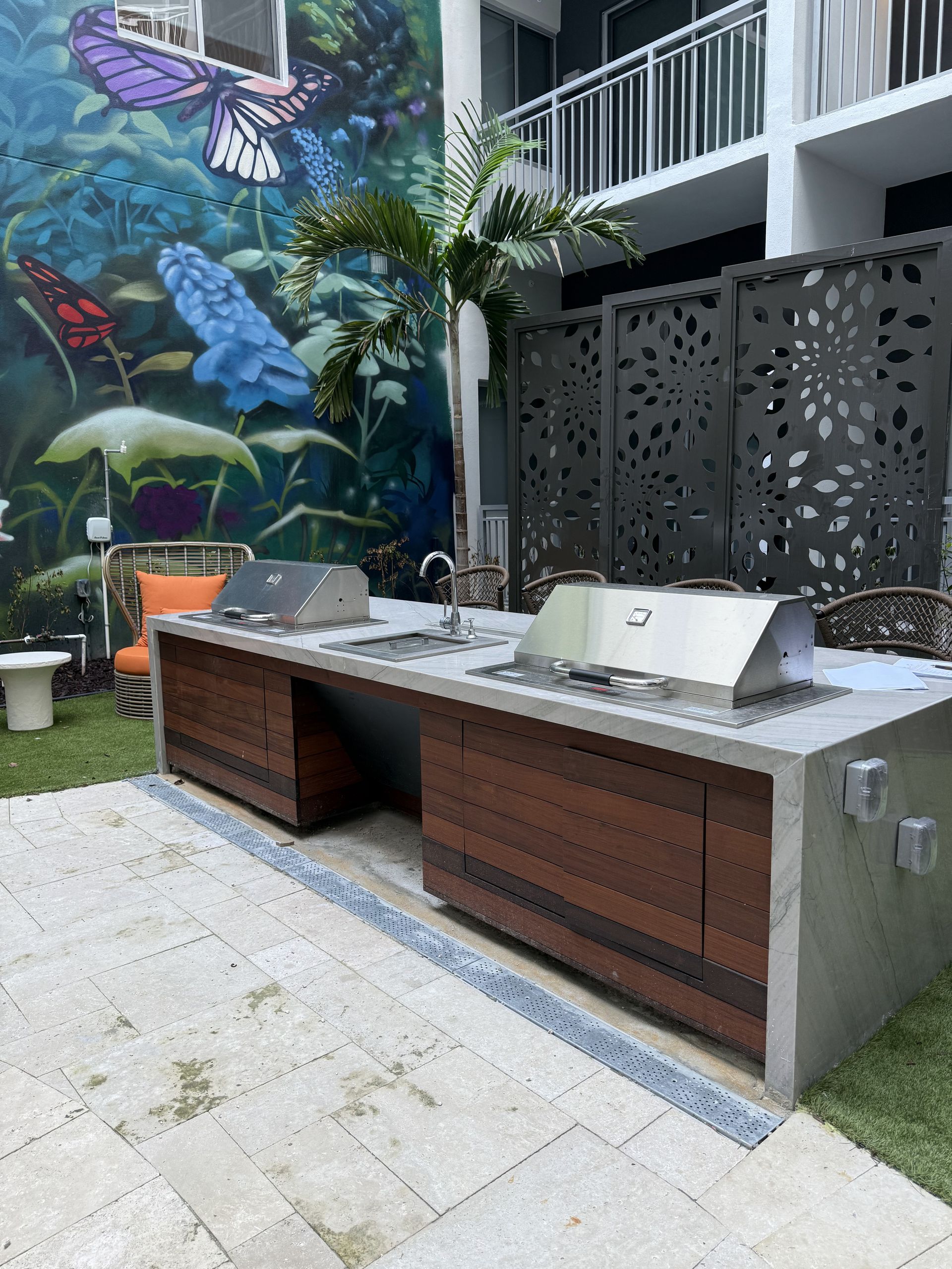 Outdoor grilling station with two grills, a sink, and wooden cabinets against a mural.