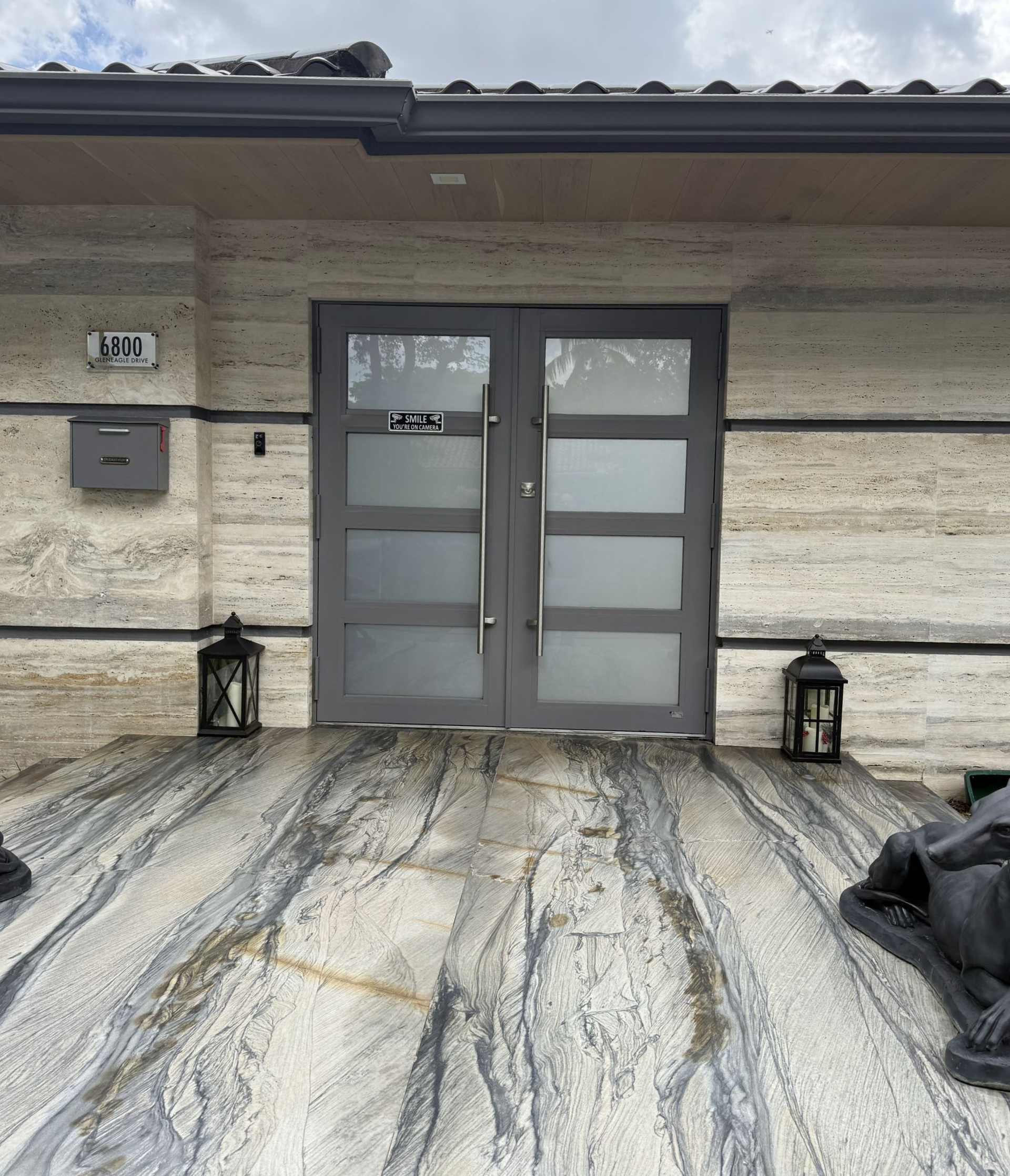 Gray double doors on a home with textured, light-colored stone facade and patterned floor. Lanterns sit on the porch.