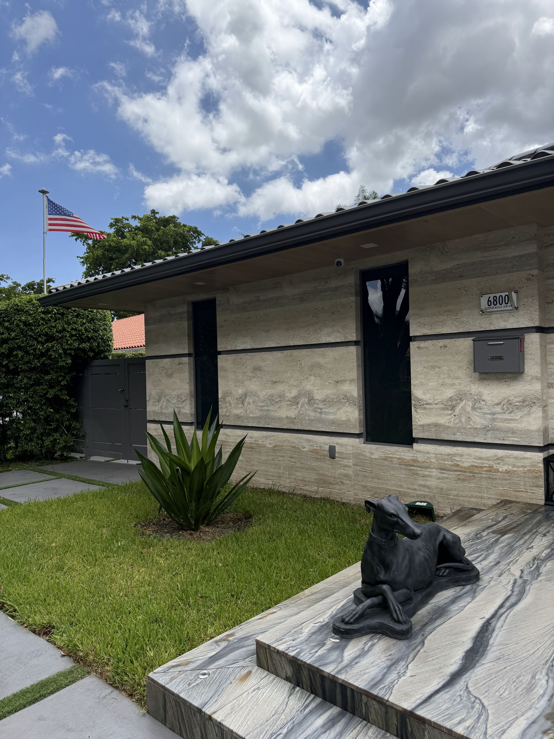 Exterior of a beige stone house with an American flag, black door, and a sculpture of a dog. Sunny, cloudy sky.