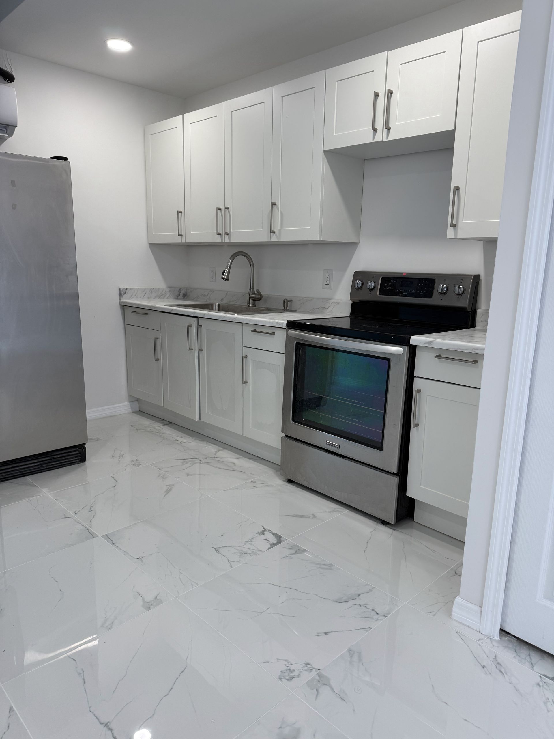Modern kitchen with white cabinets, stainless steel appliances, and white marble-look floor.