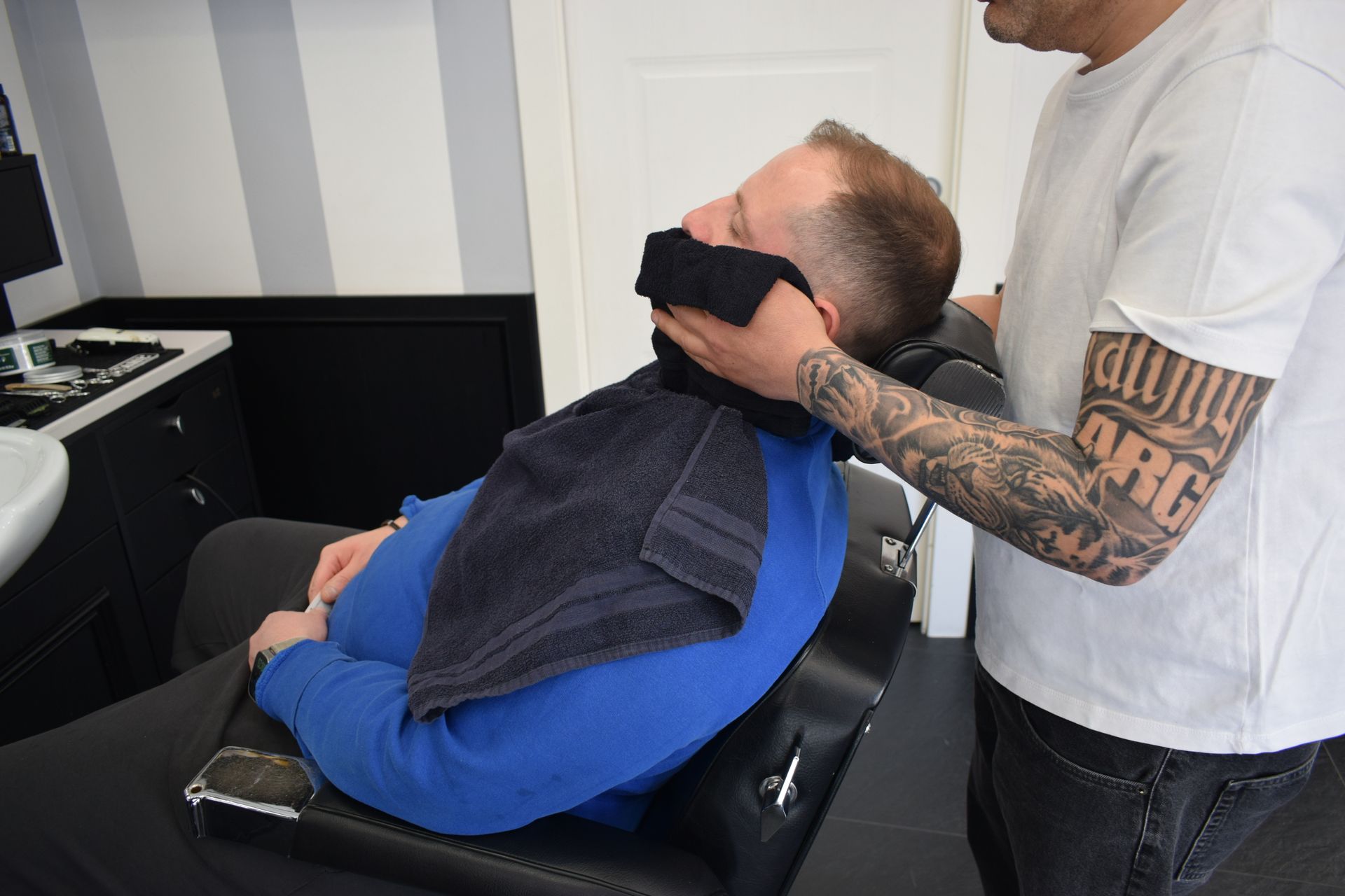 In un salone, una persona è sdraiata su una poltrona da barbiere mentre un'altra le taglia i capelli con il rasoio elettrico.