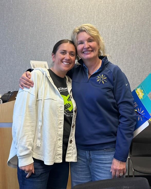 Two women, smiling and embracing in an office setting. One wears a blue jacket and jeans, the other a cream jacket over a black shirt.