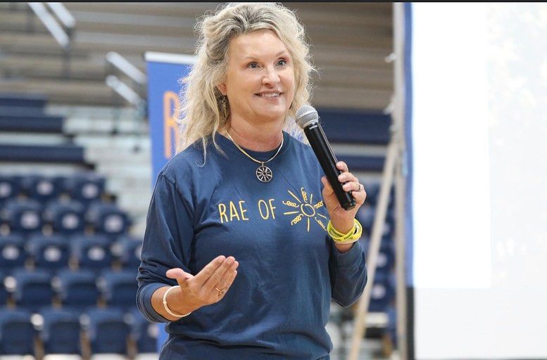 Woman with blonde hair speaking into a microphone, gesturing with her hands. She's wearing a blue shirt that says