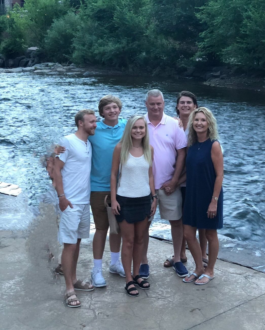Family of six smiling by a flowing river. Two men, two women, and two teenagers are posed on a stone walkway.