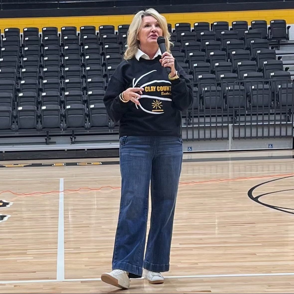 Woman with a microphone speaks on a basketball court, wearing a black sweatshirt and jeans. Empty bleachers are in the background.