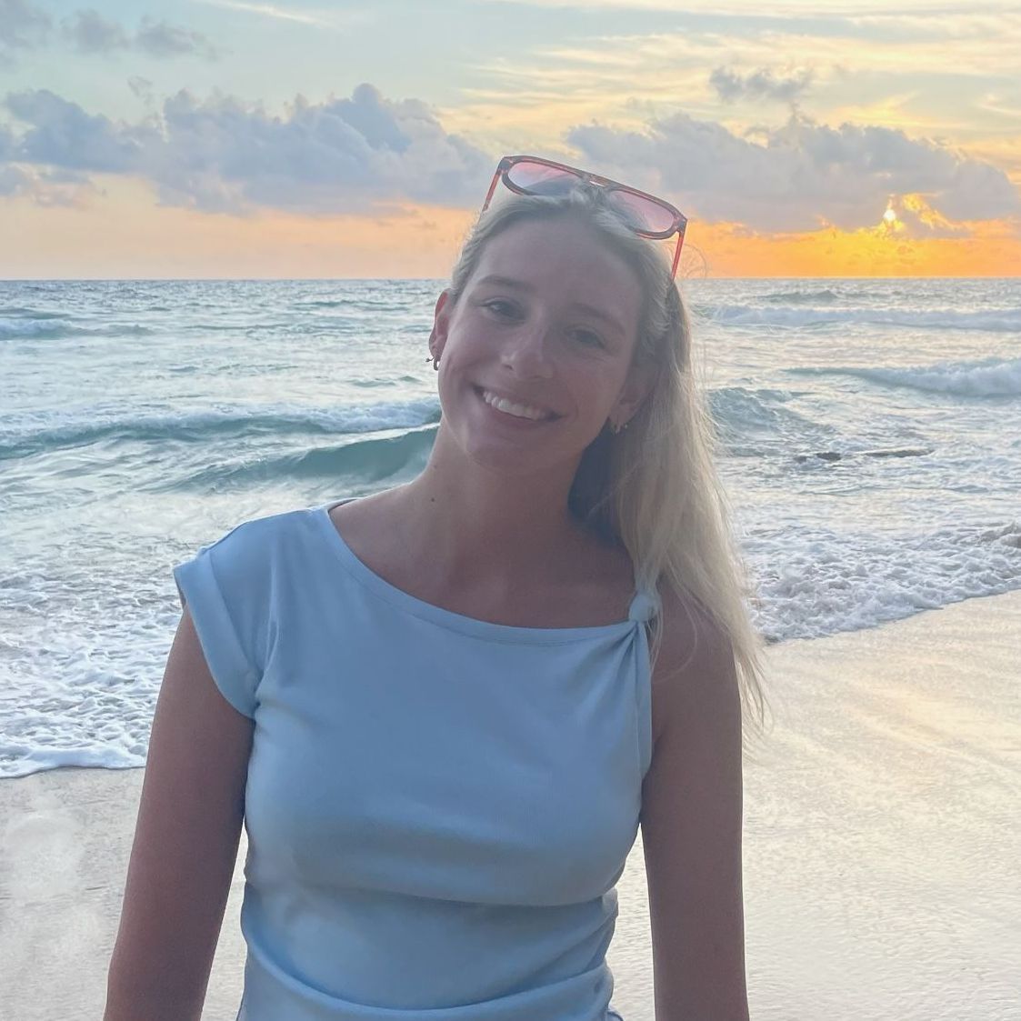 Woman smiling at the beach during sunset, wearing a light blue top and pink sunglasses on her head.