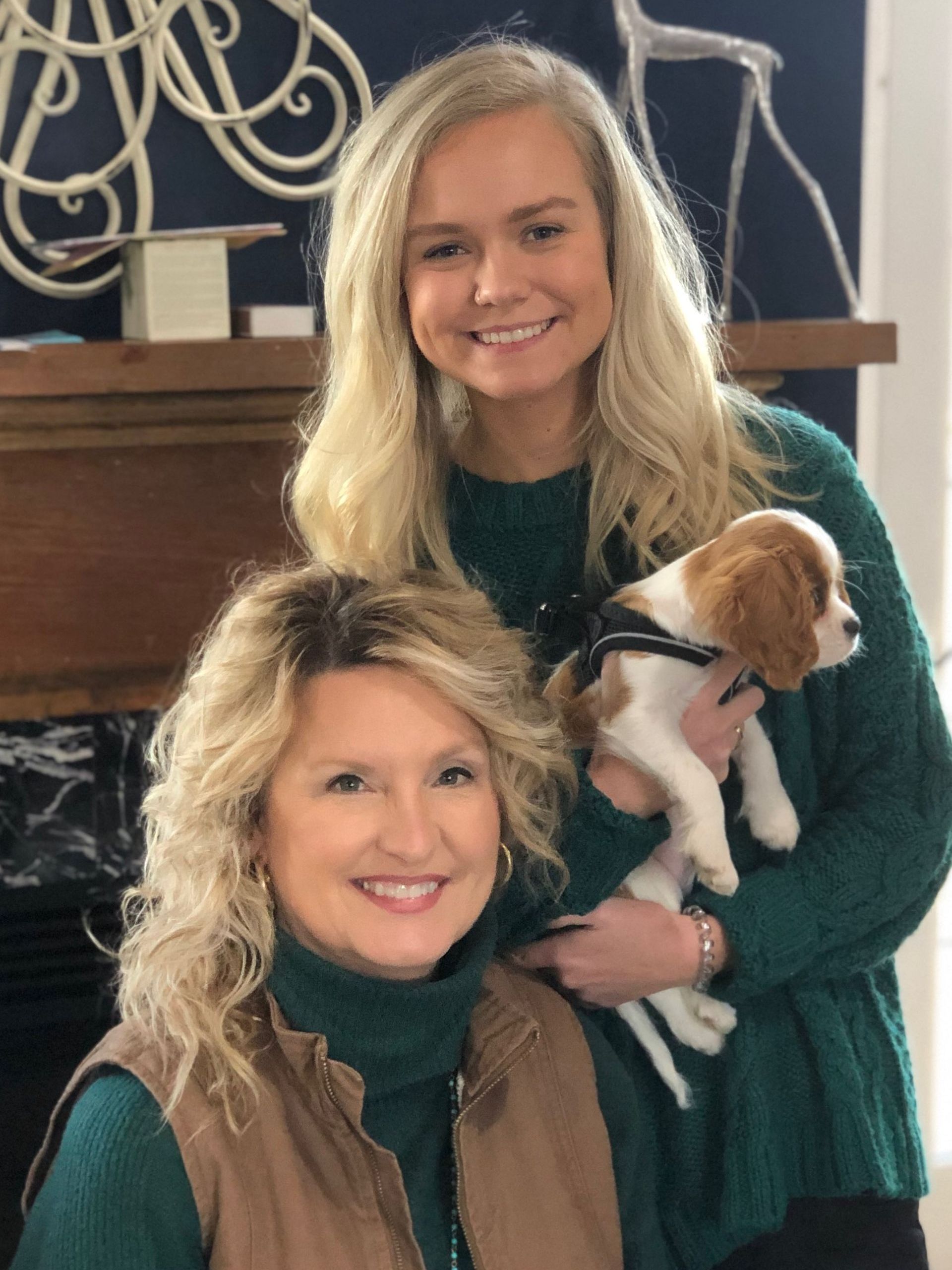 Woman and young adult woman smile while the younger woman holds a Cavalier King Charles Spaniel puppy. They are indoors with a mantel and decorations in the background.