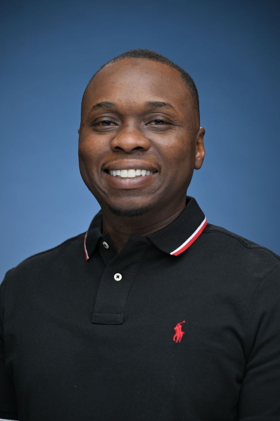 Man in black polo shirt smiling at the camera against a blue backdrop.