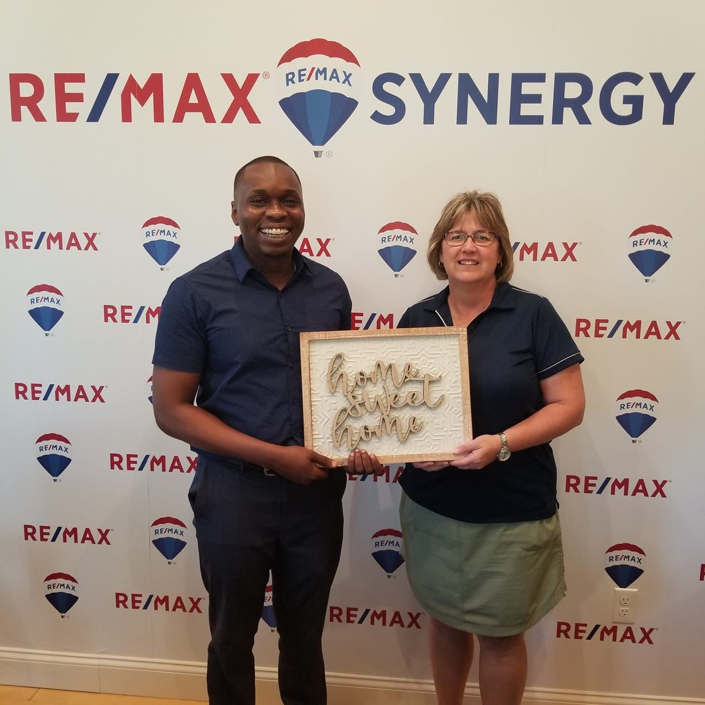 Two RE/MAX Synergy representatives holding a framed award in front of a branded backdrop