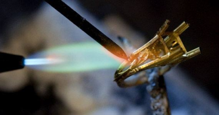 Close-up of tweezers holding a tiny golden ring base heated by a blue flame