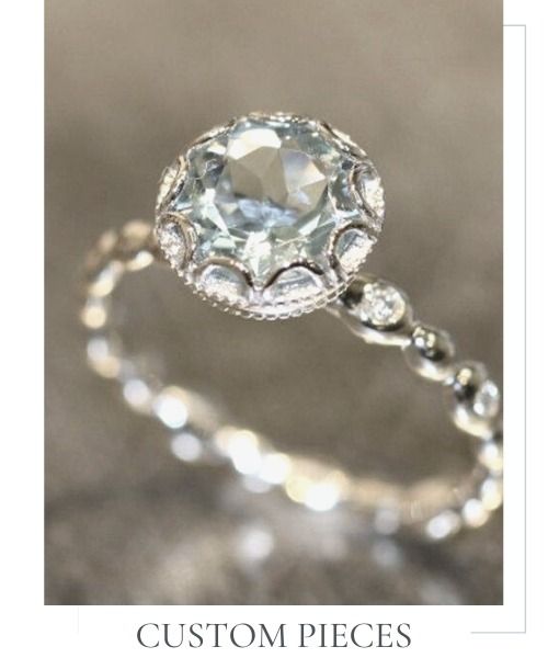 Sparkling silver ring with a round clear gemstone and smaller stones, labeled “Custom Pieces”