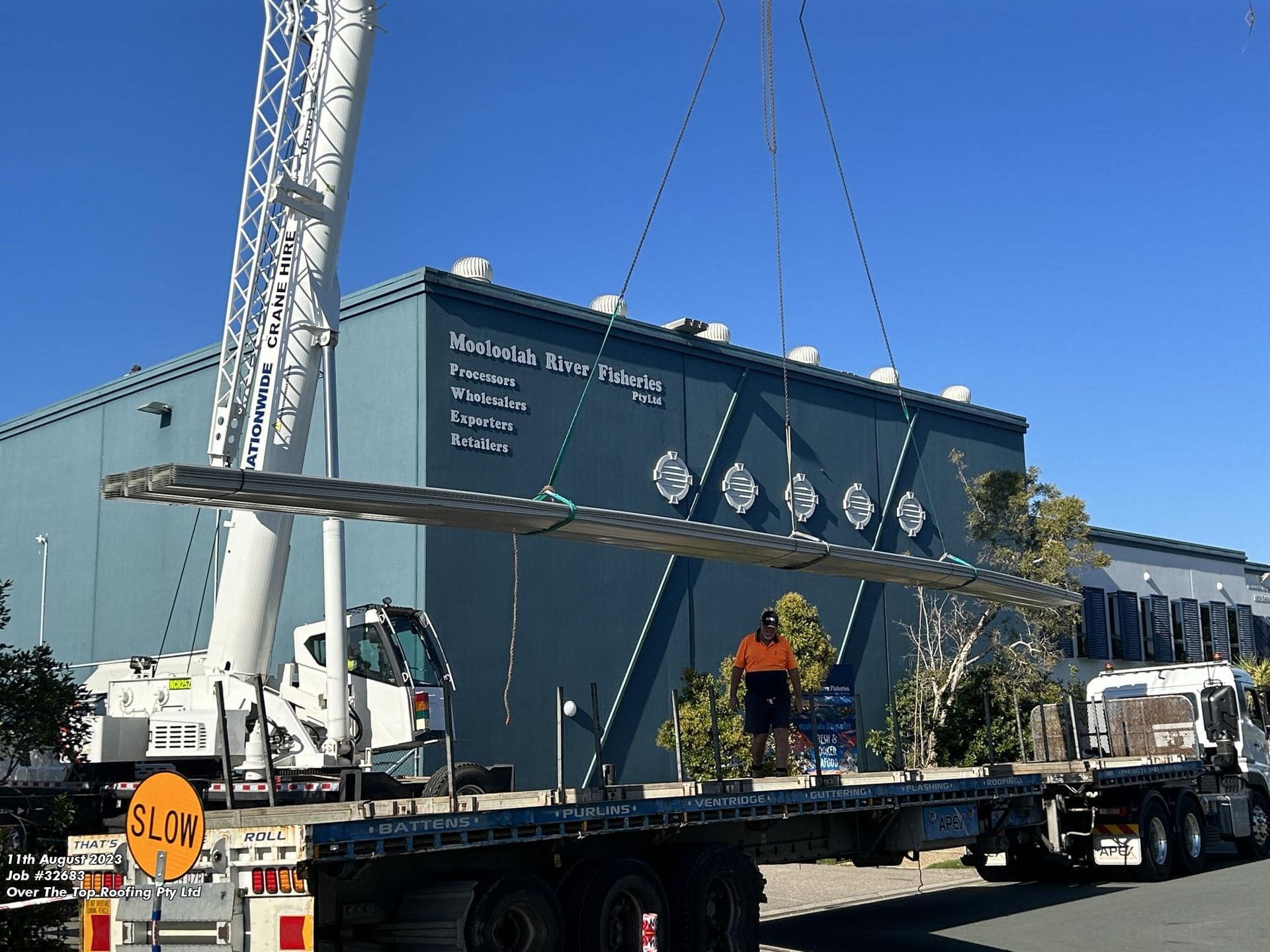 Crane Lifting Metal Beams Onto Truck in Front of a Blue Building With Sign — Over the Top Roofing Specialists in Gympie, QLD