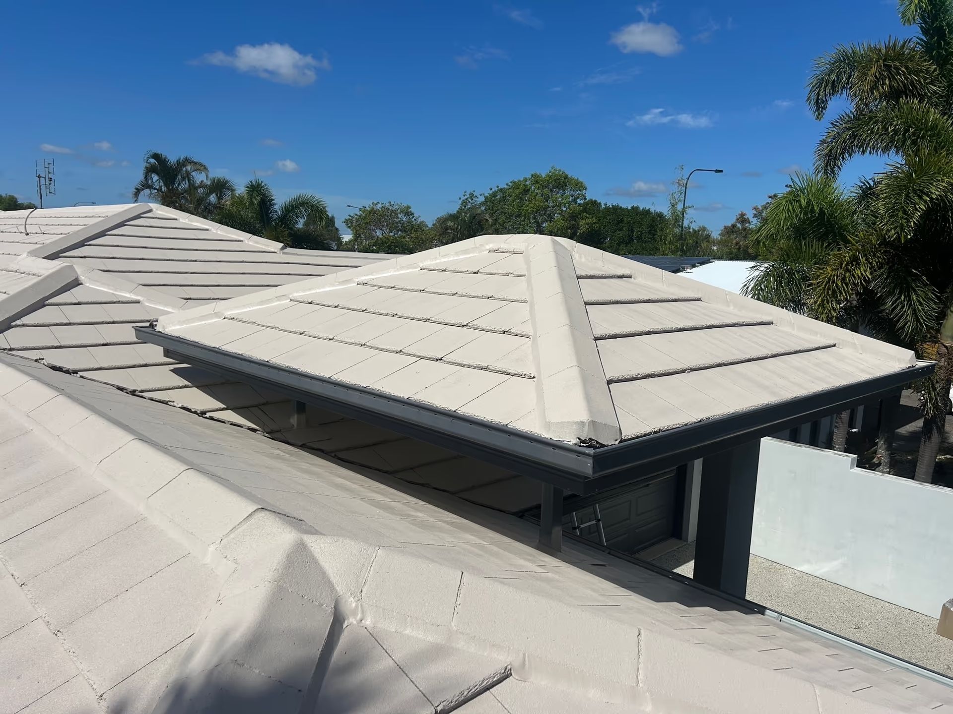 Light-colored tiled house roof under a blue sky with trees in the background